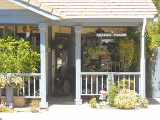 Covered porch entrance with white railing, potted plants, and a window sign that reads 'ARAGON MANOR'.
