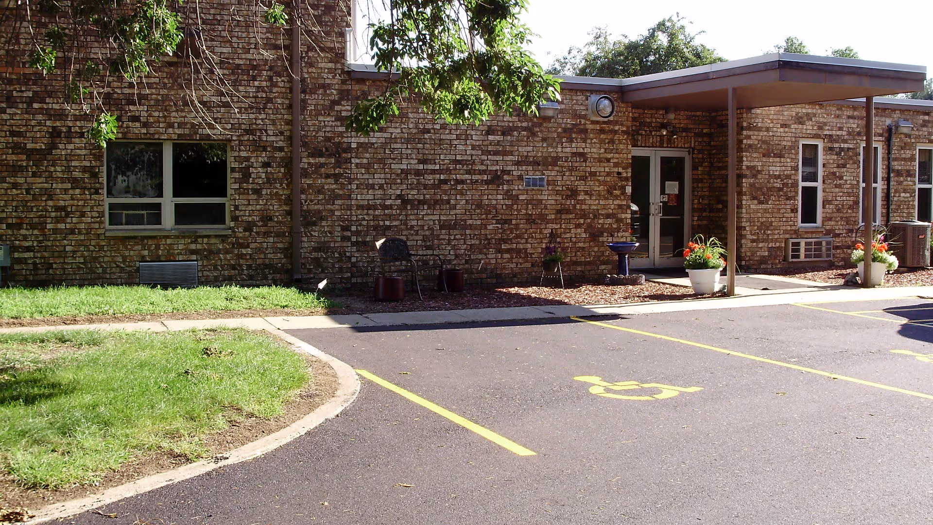 Exterior view of a single-story brick building with a covered entrance, potted plants near the door, a chair, and a handicapped parking space in front. There is a small grassy area with a tree casting shadows on the building.