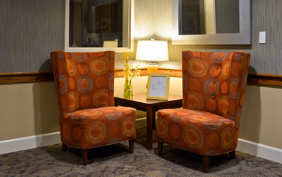 Two orange patterned armchairs facing each other with a small wooden table between them. On the table is a lamp, a small vase with yellow flowers, and a framed document. The setting is indoors with beige and gray walls and carpeted floor.