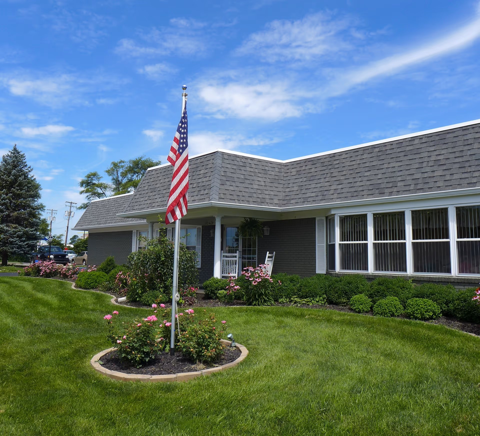 Exterior view of a single-story building with gray shingles and white trim, surrounded by well-maintained green grass and flower beds. An American flag is displayed on a flagpole in the front yard under a partly cloudy blue sky.