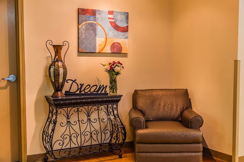 A cozy corner with a brown leather armchair next to a decorative black metal console table. On the table, there is a large vase, a glass vase with flowers, and a decorative word sign that says 'Dream'. Above the table hangs a colorful abstract painting. The walls are painted beige.