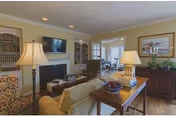 A cozy living room with a beige sofa, two table lamps, a flat-screen TV mounted above a fireplace, built-in bookshelves, and a wooden sideboard with a framed picture and plants. The room opens into a dining area with a table and chairs.