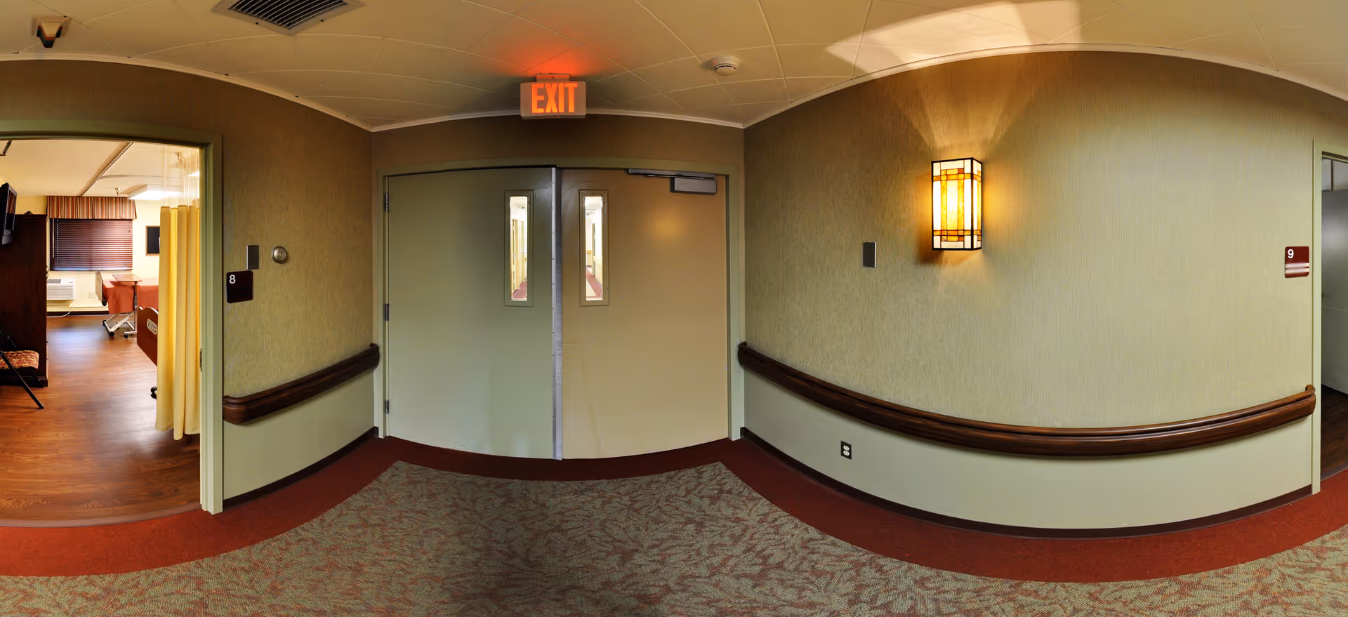 A hallway in a senior living facility with carpeted floor and beige walls. There are two doors in the center with small windows and an illuminated exit sign above them. On the left side, an open doorway reveals a room with a hospital bed, a TV mounted on the wall, and a window with blinds. The hallway has handrails along the walls and a wall-mounted light fixture on the right side.