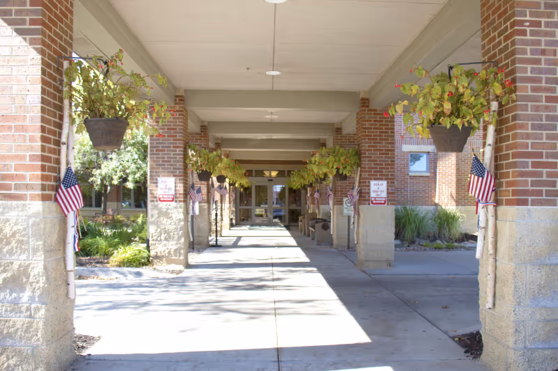 Covered entrance walkway with brick columns, hanging plants, and small American flags leading to glass doors.