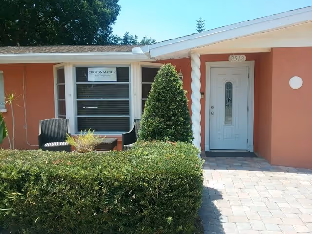 Front exterior view of a single-story building with a white door and a window displaying a sign that reads 'Croton Manor'. There are two black chairs and a small table outside near the window, surrounded by green bushes and a small tree. The building is painted in a reddish color with a white roof trim.