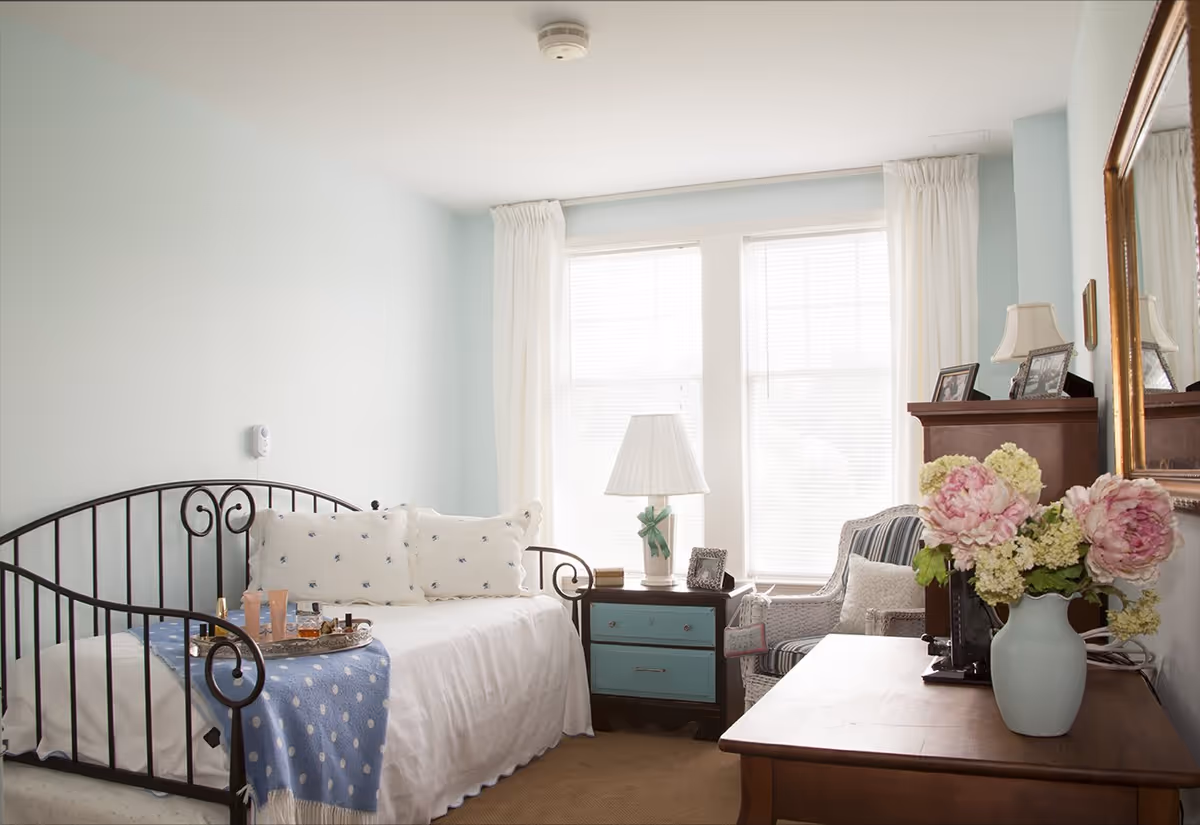 A bright and cozy bedroom with a black metal daybed adorned with white pillows and a blue polka dot throw blanket. A small wooden nightstand with blue drawers holds a white lamp and picture frames. Next to the nightstand is a striped armchair with a cushion. A wooden dresser with a large mirror above it displays a vase of pink and white flowers. The room has light blue walls and large windows with white curtains letting in natural light.