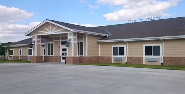 Exterior view of a single-story building with beige siding and brick accents, featuring a peaked roof and a covered entrance. The building is set against a partly cloudy sky with a concrete driveway in front.