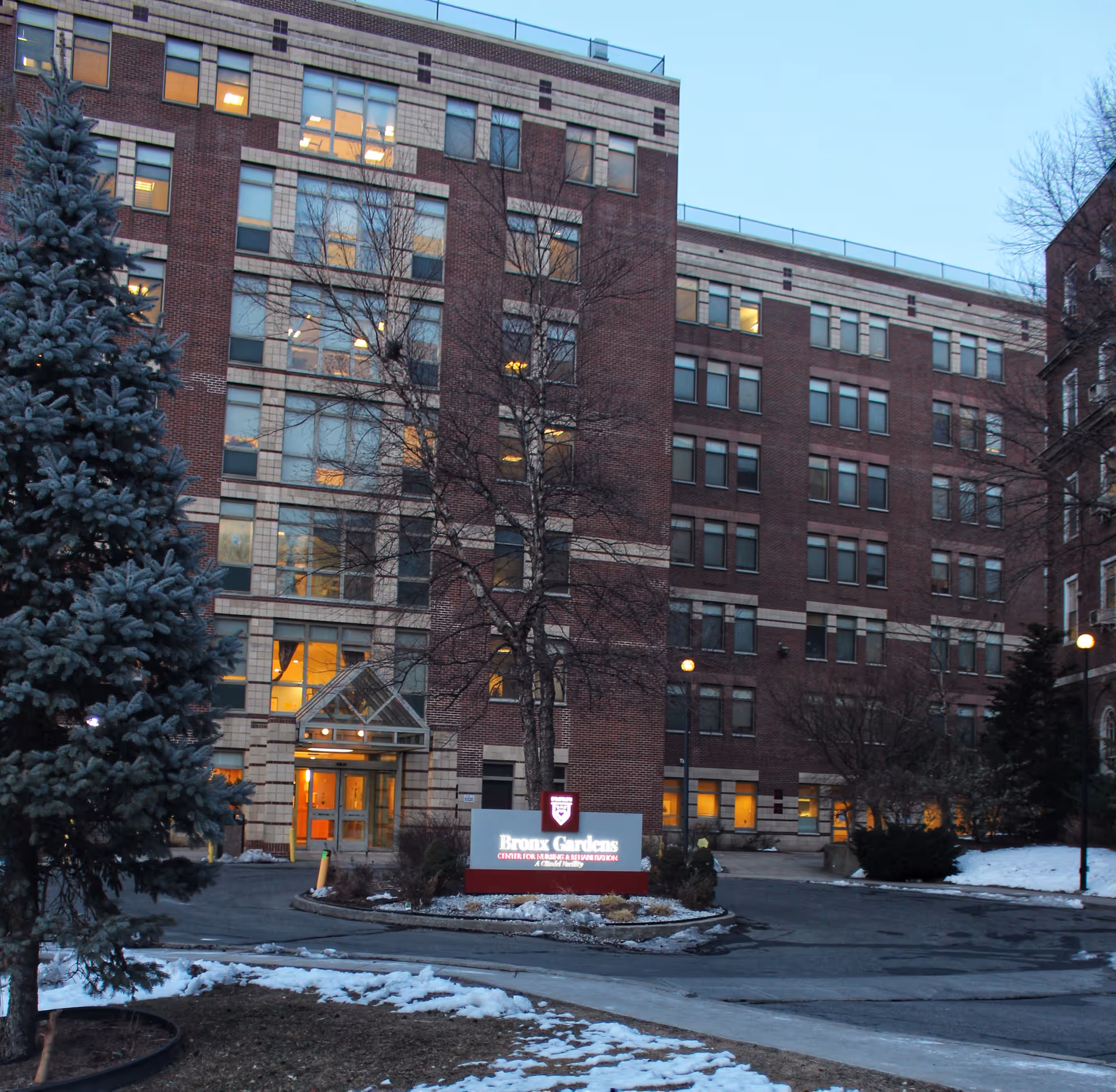 Exterior front of a multi-story brick nursing and rehabilitation building with a lit main entrance, a 'Bronx Gardens' sign, and patches of snow on the ground.
