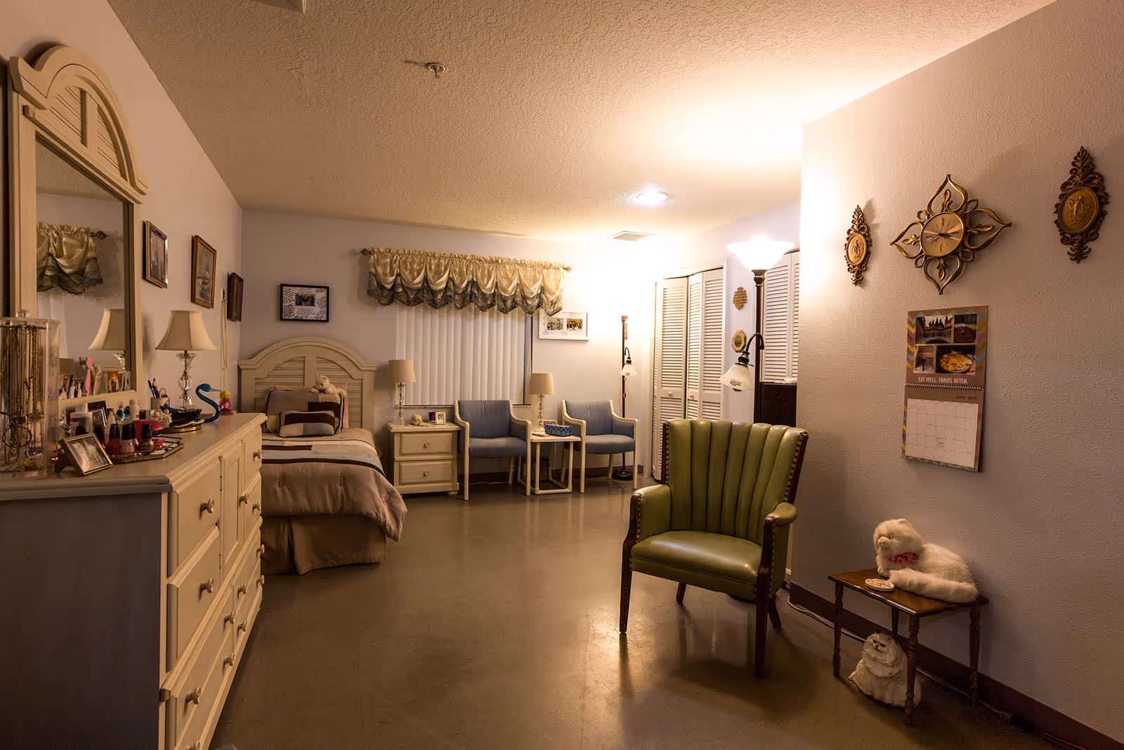 A cozy bedroom in an assisted living facility featuring a bed with pillows and a stuffed animal, a dresser with a large mirror, two blue chairs with a small table between them, a green armchair, a floor lamp, and decorative wall hangings including a clock and framed pictures. The room has soft lighting and a calendar on the wall.