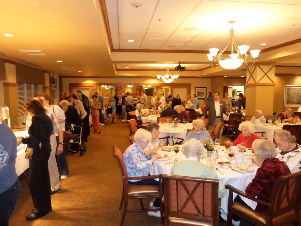 A dining room in a senior living facility with elderly residents seated at round tables enjoying a meal. Other people are standing in line at a buffet table on the left side of the room. The room is warmly lit with chandeliers and has a cozy, welcoming atmosphere.