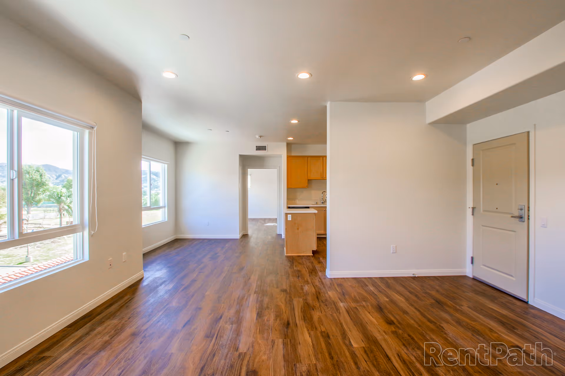 Empty interior space with wooden flooring, white walls, recessed ceiling lights, large windows showing an outdoor view with trees and mountains, a small kitchen area with wooden cabinets, and a closed door on the right side.