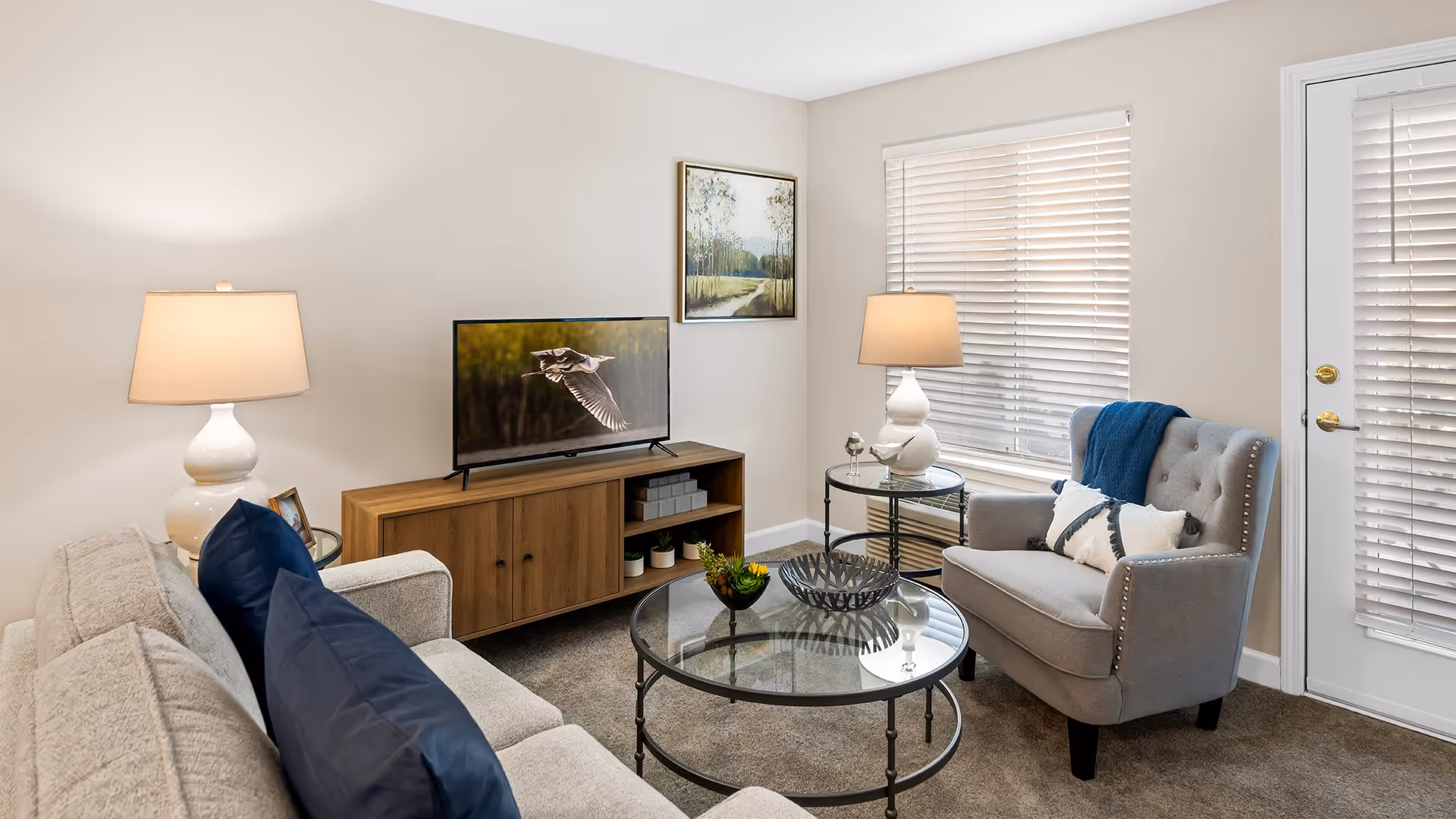 A cozy living room with a beige sofa adorned with dark blue pillows, a gray armchair with a white and blue pillow and a blue throw blanket, two white table lamps on glass side tables, a wooden TV stand with a flat-screen TV displaying a bird in flight, a round glass coffee table with decorative items, a window with white blinds, and a door with white blinds.