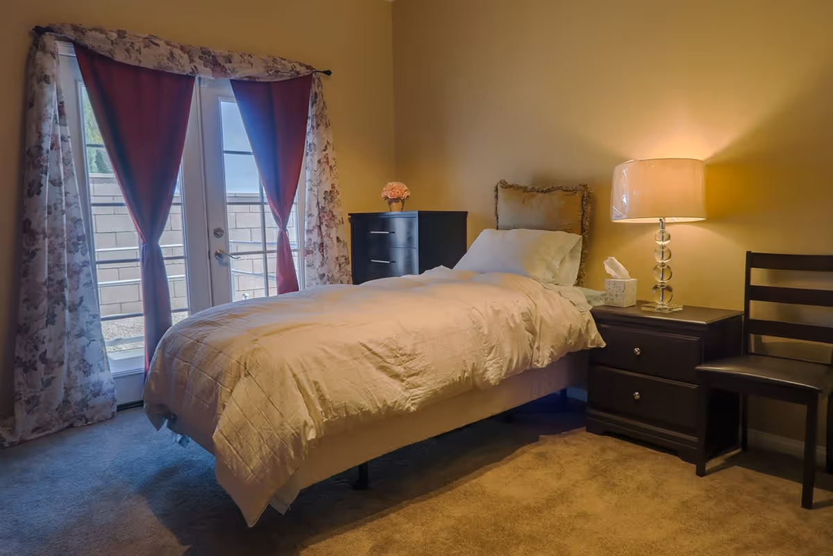 A cozy bedroom with a single bed covered in a beige comforter and a pillow. Next to the bed is a dark wooden nightstand with a lamp and a box of tissues. There is a dark wooden chair beside the nightstand. In the background, there is a dresser with a small flower vase on top. The room has beige walls and carpeted floor. French doors with floral and red curtains lead outside to a small patio area enclosed by a brick wall.