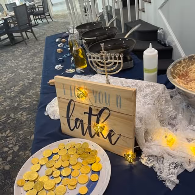 A table set up with Hanukkah-themed decorations including a menorah, a wooden sign that says 'I love you a latke', a plate filled with chocolate gold coins, and some cooking pans and utensils in the background.