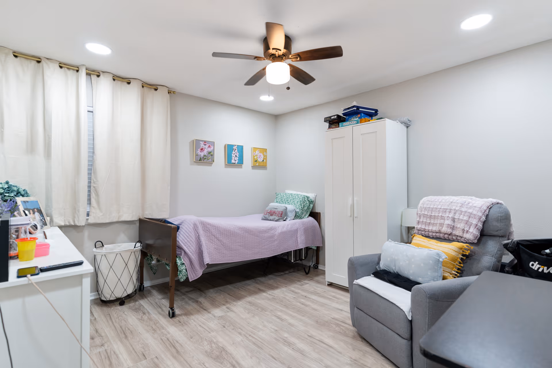 A cozy bedroom with a single bed covered in a lavender blanket and green pillows, a white wardrobe with board games on top, a gray armchair with multiple cushions and a pink throw blanket, light wood flooring, white walls with three small floral paintings, a ceiling fan with light, and a window with cream-colored curtains.