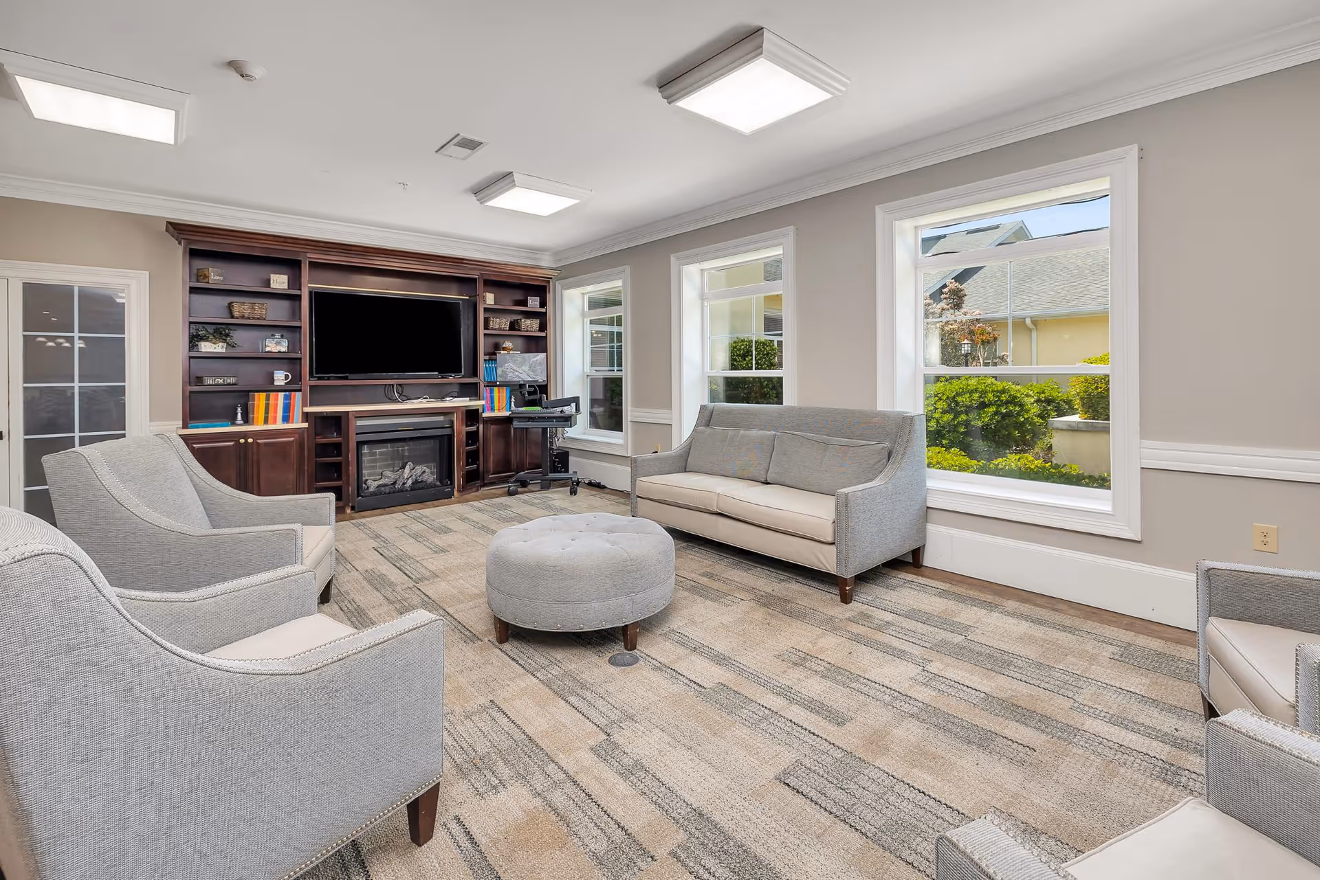 A bright and spacious living room with beige walls and large windows letting in natural light. The room features a gray sofa, two matching armchairs, a round gray ottoman, and a dark wooden entertainment center with a flat-screen TV and a fireplace. There is also a small desk with a computer chair near the windows.