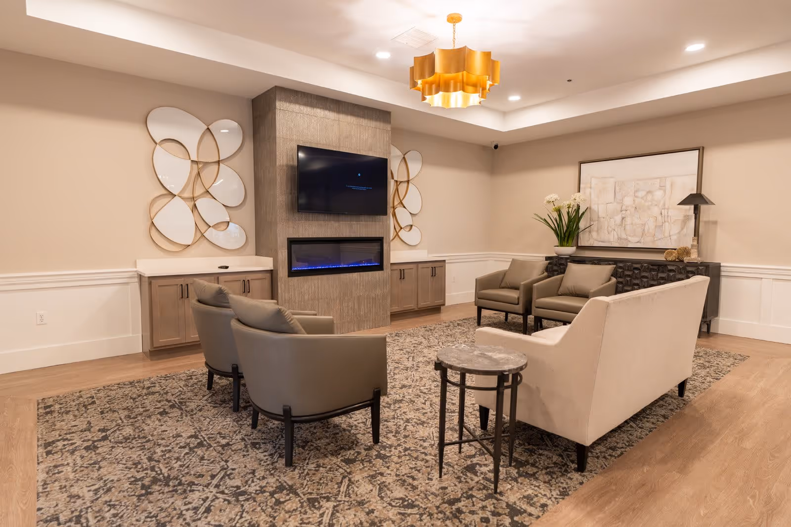 A cozy living room area in a senior living facility featuring a beige sofa, two gray armchairs, and two additional chairs arranged around a patterned rug. The room has a modern electric fireplace with a mounted flat-screen TV above it, decorative wall art on either side, a side table, and a console table with a lamp and a vase of flowers. The ceiling has recessed lighting and a stylish gold chandelier.