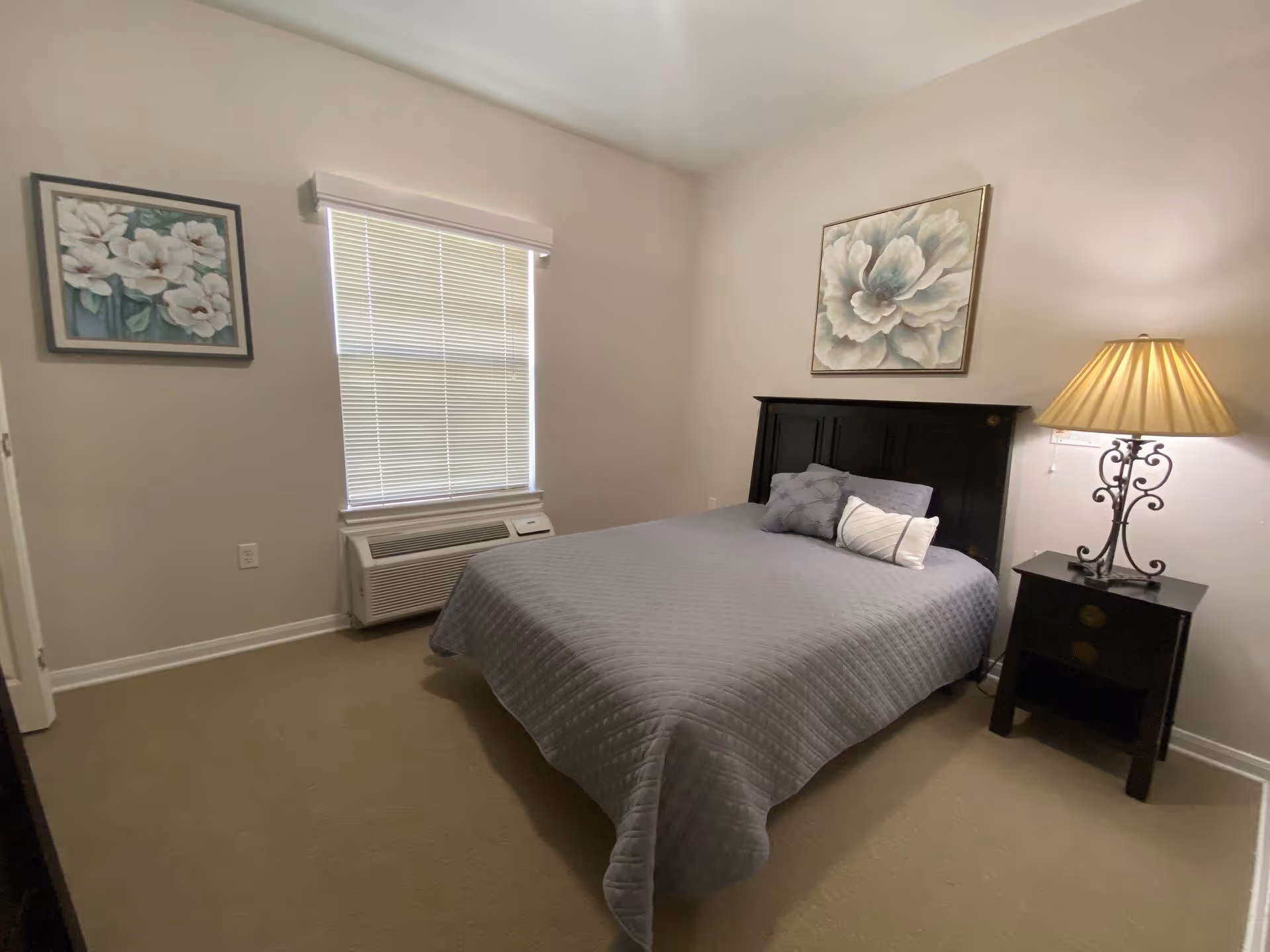 A bedroom with a neatly made bed covered with a gray quilt and three decorative pillows. There is a black headboard and a matching black nightstand with a lamp on it. Two floral paintings hang on the walls, one above the bed and one next to a window with closed blinds. The room has beige carpet and light-colored walls.