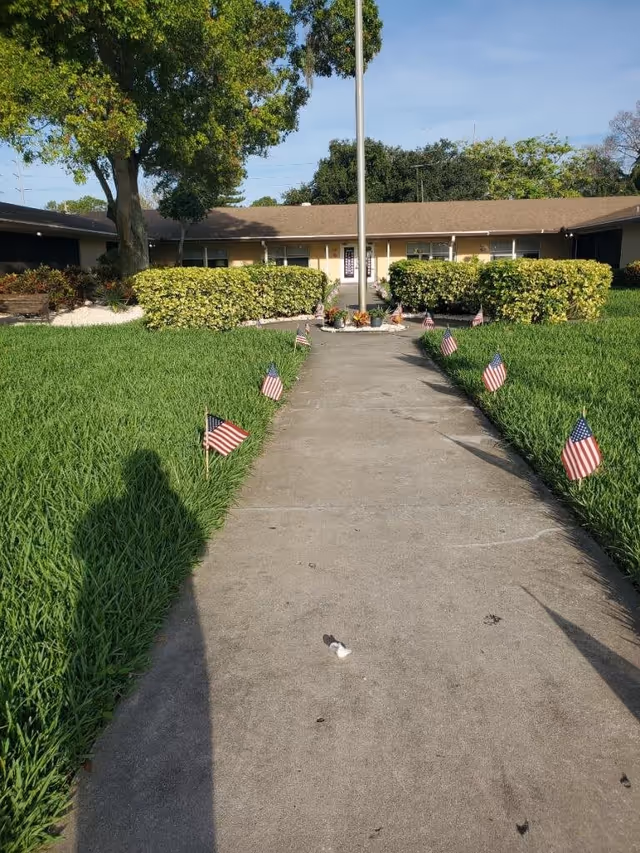 A concrete walkway lined with small American flags on both sides leads to a single-story building with a brown roof and beige walls. There is a flagpole in the center near the building entrance, surrounded by bushes and greenery. Trees and a clear blue sky are visible in the background.