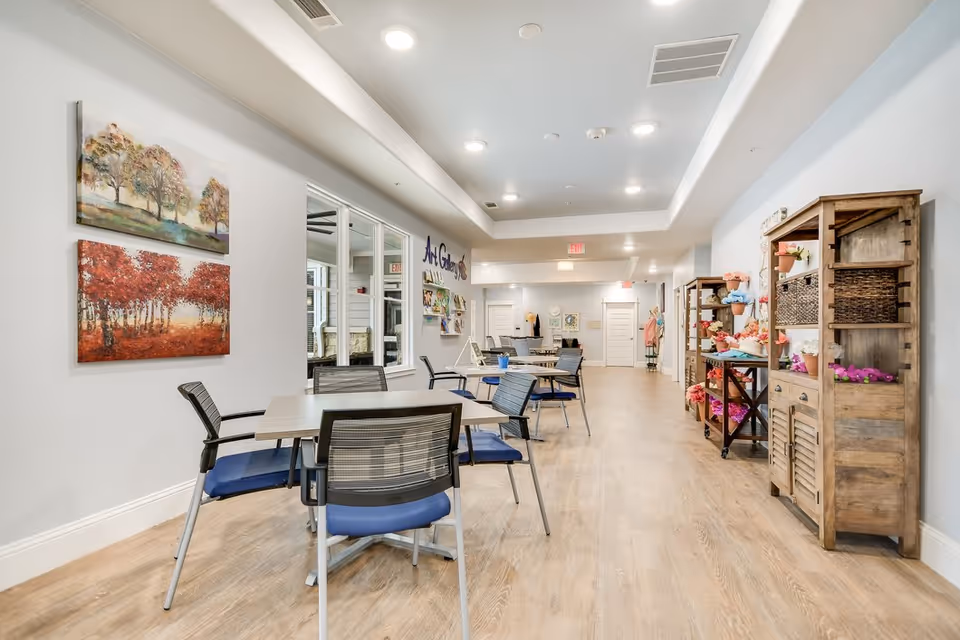 Bright communal activity room with tables and chairs, artwork on the walls, and wooden shelving displaying decorative items.