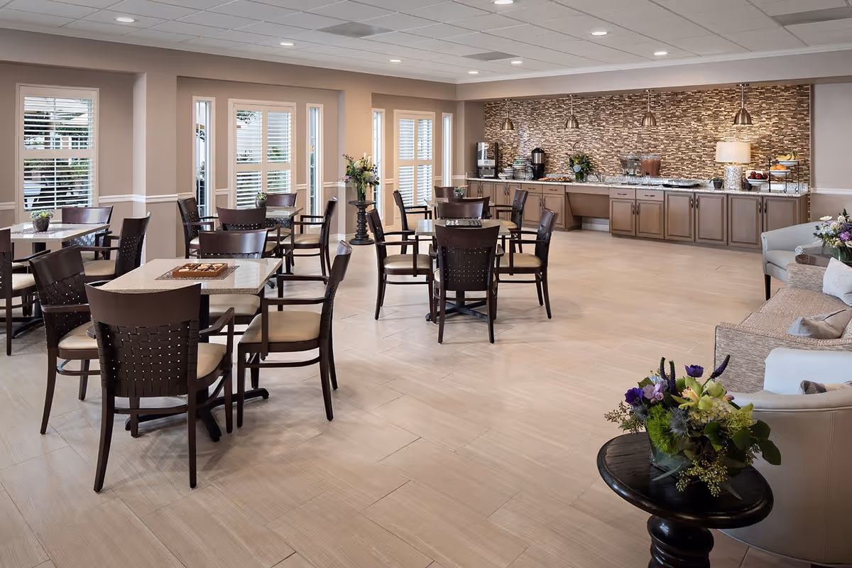 A bright and spacious dining area in a senior living facility with multiple tables and chairs arranged neatly. The room features large windows with white blinds, a buffet counter with coffee and beverage dispensers, and decorative pendant lights hanging from the ceiling. There are also comfortable seating areas with sofas and armchairs, and floral arrangements on tables adding a touch of color.
