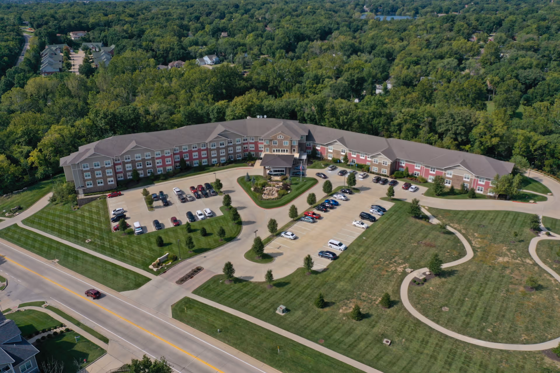 Aerial view of Cedarhurst of Columbia, a large senior living facility surrounded by green trees and lawns. The building is U-shaped with a parking lot in front, several cars parked, and a driveway leading to the entrance. There are walking paths and landscaped areas around the property.