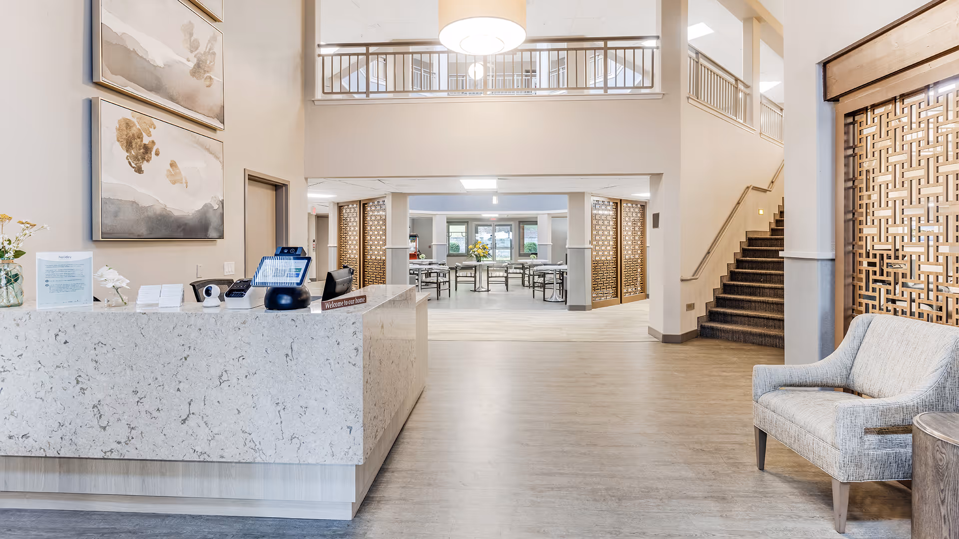 Reception area of Holiday Columbia Pines facility with a marble front desk on the left, a gray upholstered chair on the right, decorative wall art above the desk, and a staircase leading upstairs. In the background, there is a spacious common area with tables and chairs, illuminated by natural light from windows.