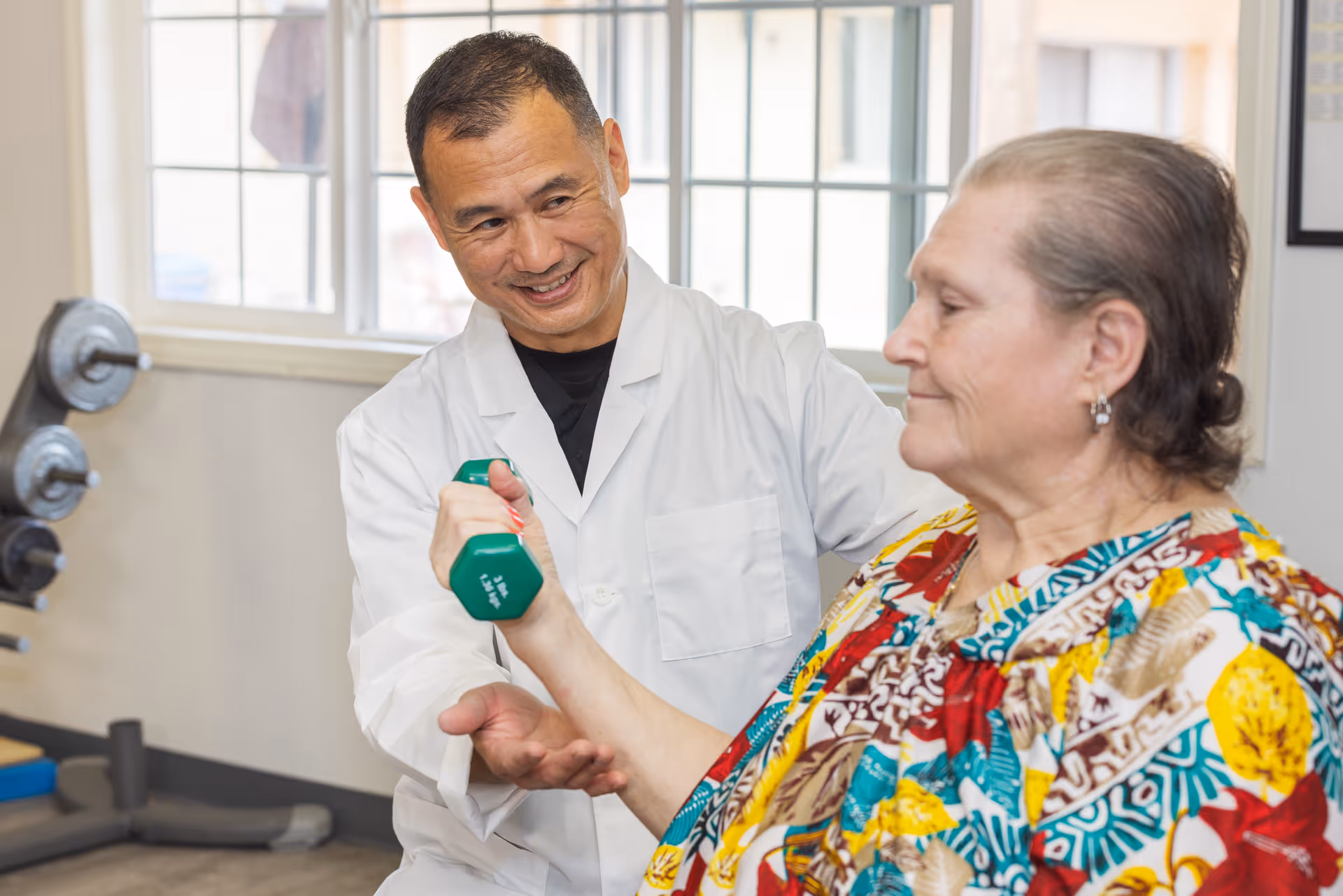 A healthcare professional in a white coat assists an elderly woman with lifting a green dumbbell in a well-lit room with large windows.