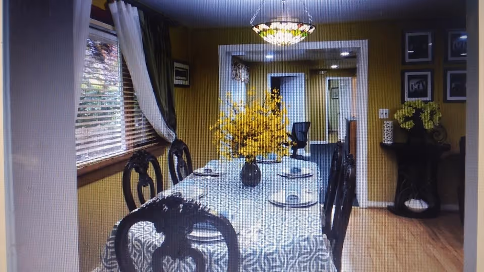 A dining room with a long table covered by a patterned tablecloth, set with plates and napkins. There are six dark wooden chairs around the table. A vase with bright yellow flowers is placed in the center of the table. The room has yellow walls, a window with white and dark green curtains on the left, and a decorative side table with more yellow flowers and framed pictures on the right wall. A chandelier hangs above the table, and a hallway is visible in the background.