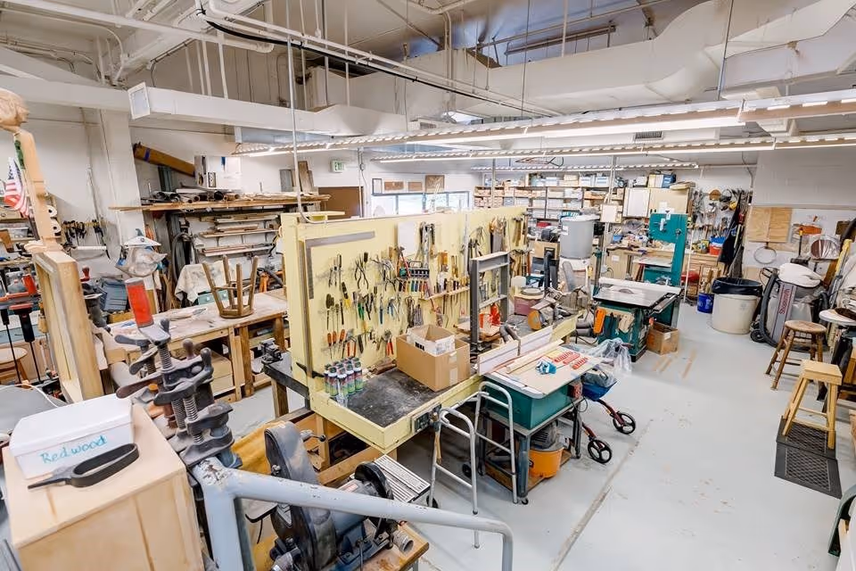A spacious workshop filled with various woodworking tools and equipment. There are workbenches, hand tools hanging on a pegboard, power tools, stools, and shelves stocked with materials and supplies. The room is well-lit with overhead fluorescent lights and has a high ceiling with exposed pipes and ducts.