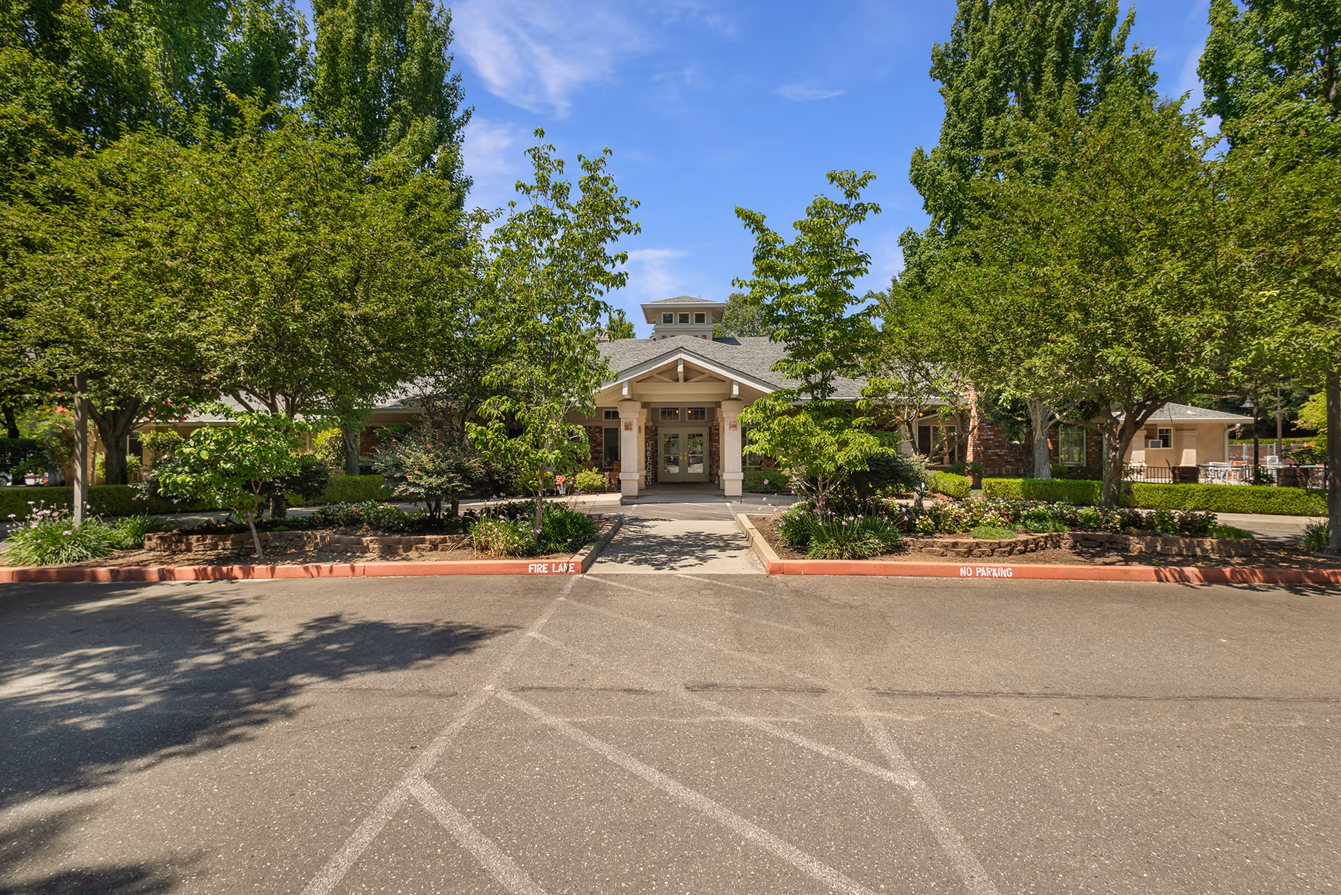 Front exterior view of Courtyard At Little Chico Creek facility with a paved driveway, landscaped garden with trees and shrubs, and a covered entrance under a clear blue sky.