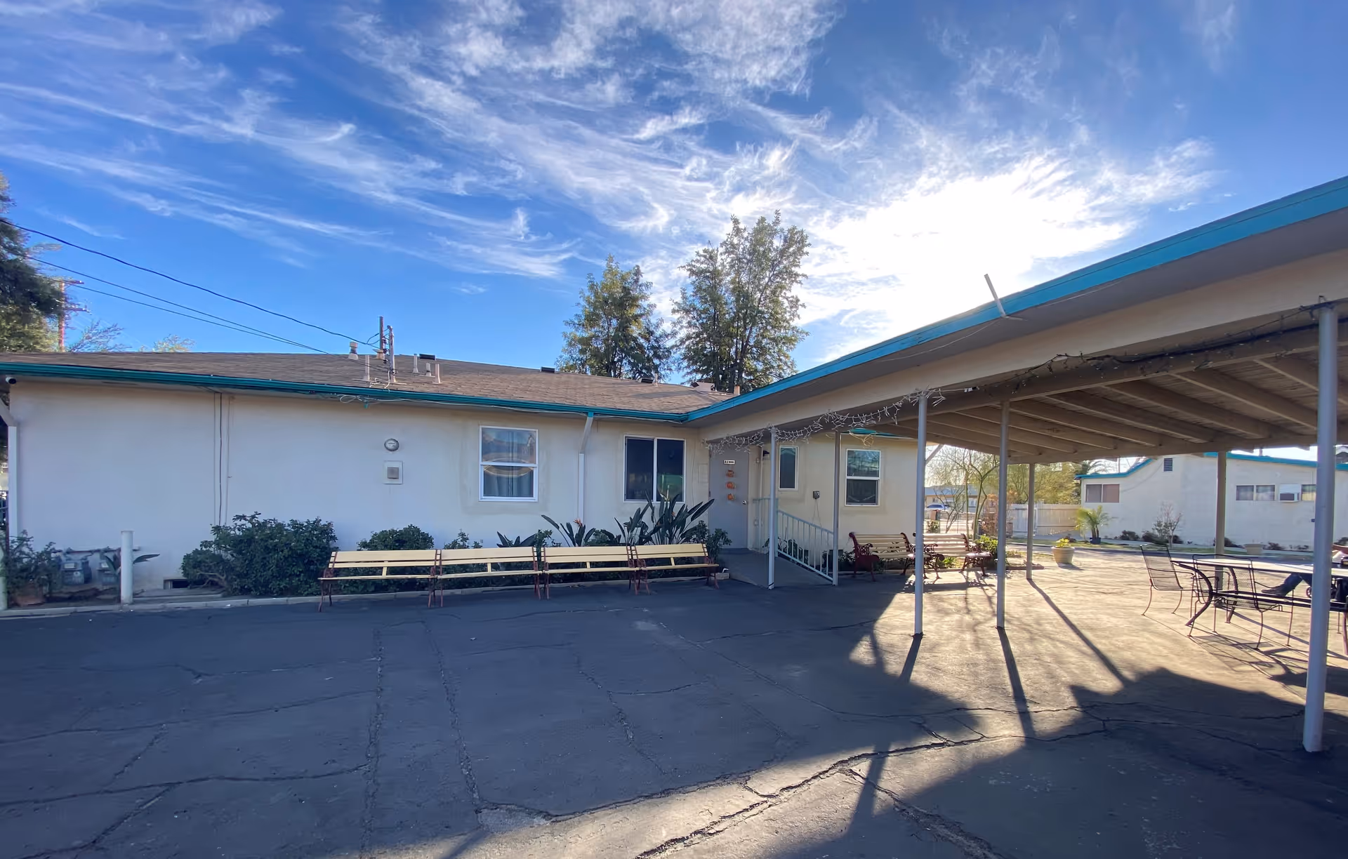 Outdoor view of a single-story building with a covered patio area supported by metal poles. There are benches along the building wall and patio, some plants near the building, and a clear blue sky with wispy clouds above.