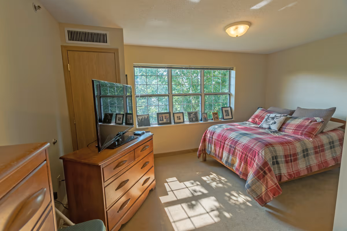 Sunlit bedroom with a plaid-covered bed, wooden dresser with a TV, and a window sill lined with framed photos.