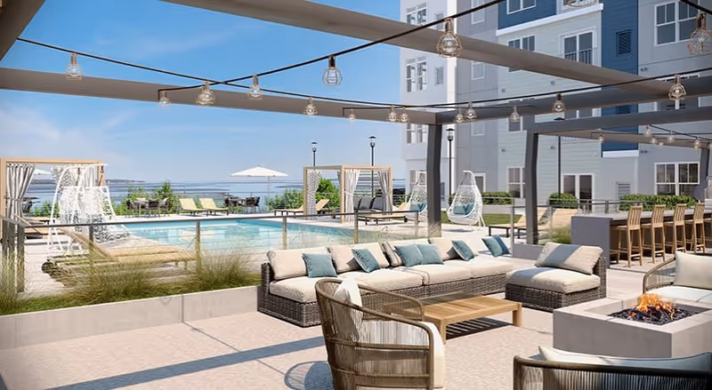 Outdoor seating area at a senior living facility featuring a large sectional sofa with cushions, a fire pit, hanging string lights, and a pool with lounge chairs and cabanas in the background under a clear blue sky.