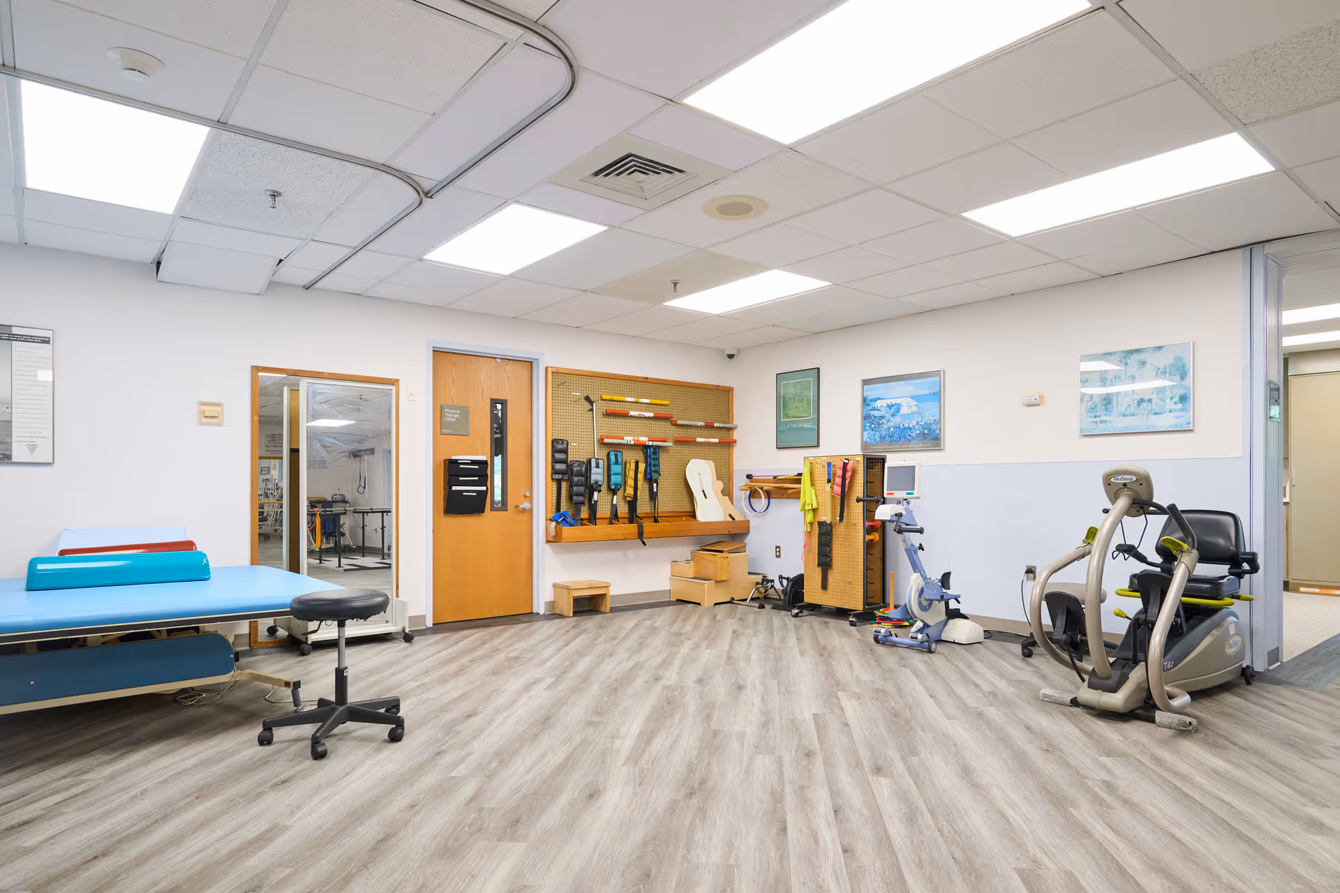 A rehabilitation therapy room with exercise equipment including a stationary bike, therapy tables with cushions, a stool, and various therapy tools mounted on the wall. The room has light wood flooring, white walls with a light blue lower half, and fluorescent ceiling lights. There are framed pictures on the walls and a wooden door with a window.