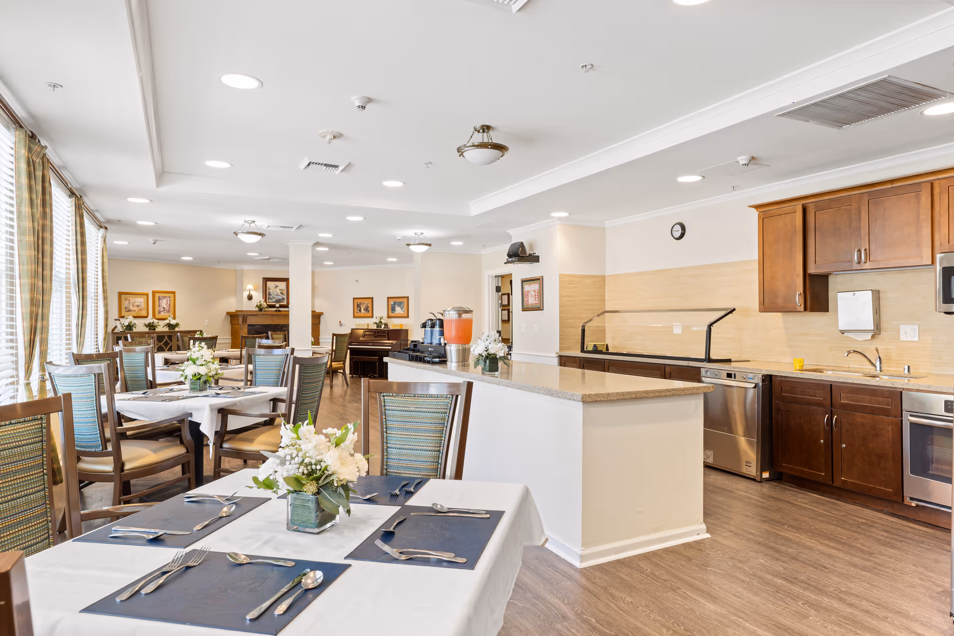 A bright dining room with several tables set with white tablecloths, blue placemats, silverware, and floral centerpieces. The room features large windows with striped curtains, wooden chairs with green cushions, and a kitchen area with wooden cabinets, stainless steel appliances, and a countertop with a beverage dispenser. The space has a warm and inviting atmosphere with framed artwork on the walls and a piano in the background.
