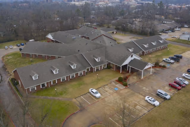 Aerial view of a large single-story brick building with multiple roof sections and dormer windows, surrounded by parking lots with several cars parked. The building is situated in a suburban area with trees and other buildings in the background.