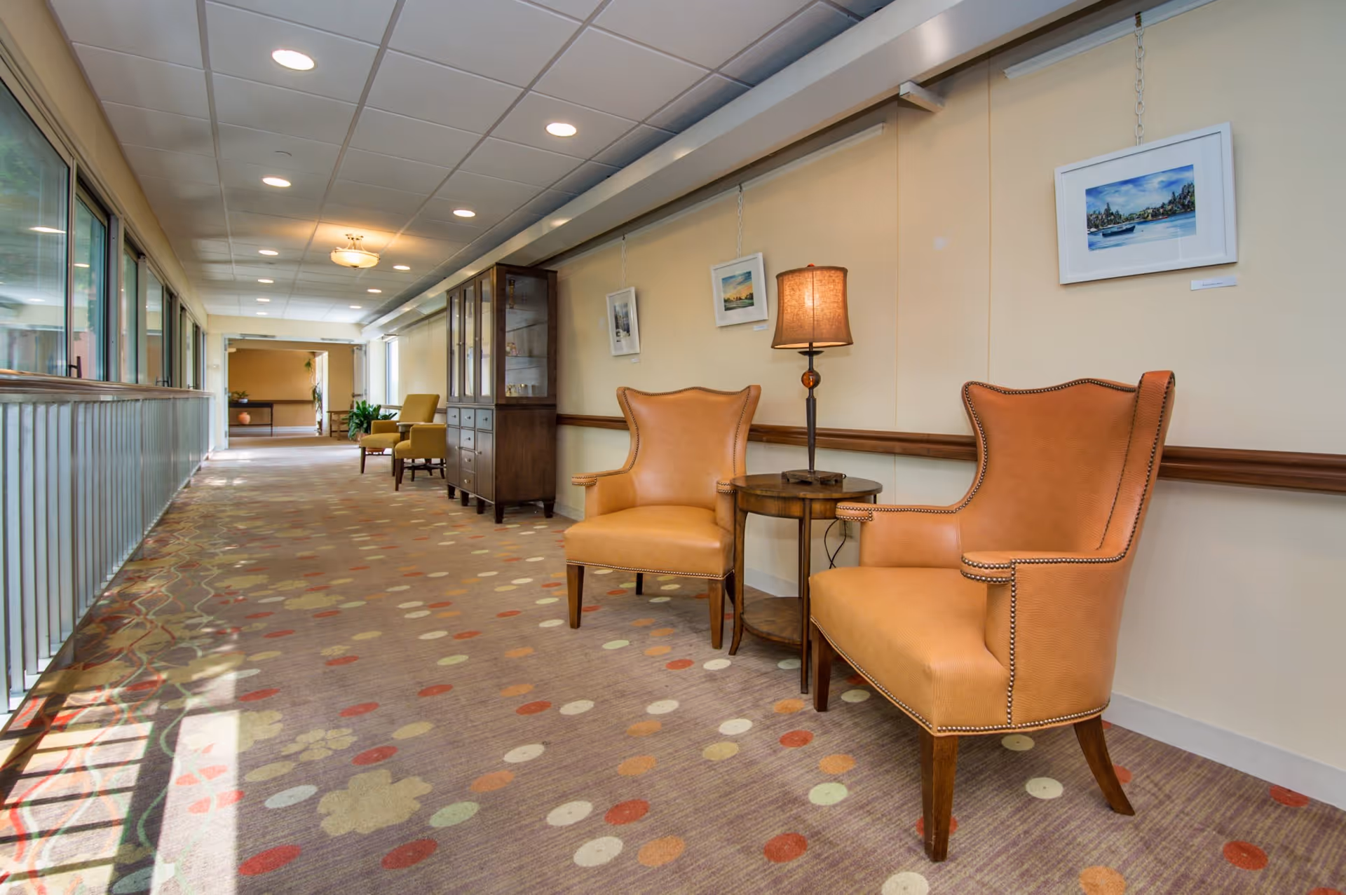 A bright hallway in a senior living facility with large windows on the left side allowing natural light to fill the space. On the right side, there are two tan leather armchairs with nailhead trim positioned around a small round wooden table with a lamp. Above the chairs, three framed paintings are hung on the wall. Further down the hallway, additional seating and a wooden cabinet are visible. The carpet has a pattern of colorful dots and shapes.