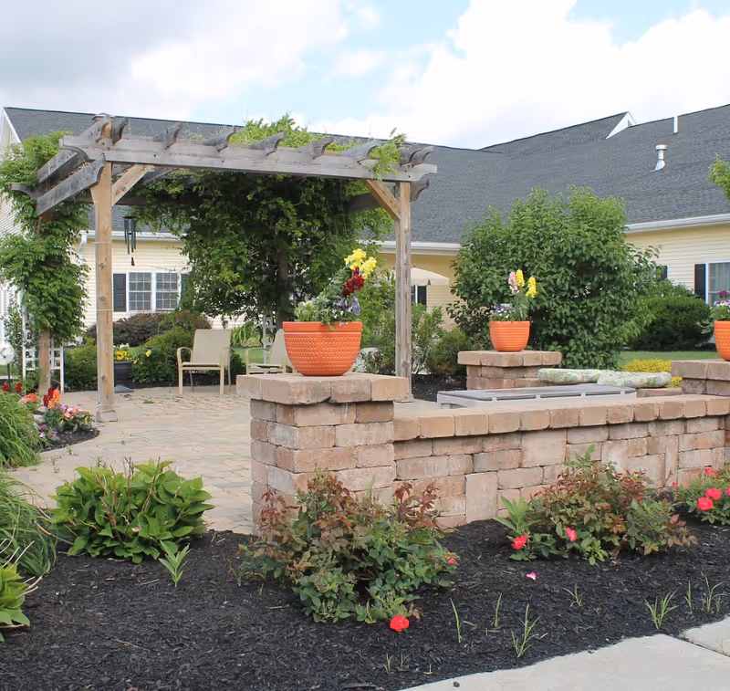 Outdoor garden area at Rochester Presbyterian Home (Cottage Grove) featuring a wooden pergola with climbing plants, patio chairs, a stone wall with orange flower pots containing colorful flowers, and well-maintained landscaping with bushes and flowers.