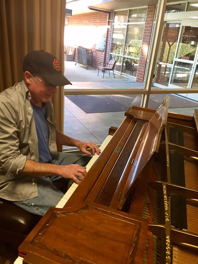 A man wearing a cap and glasses is playing a wooden piano indoors near a large window showing an outdoor patio area with chairs and a brick wall.