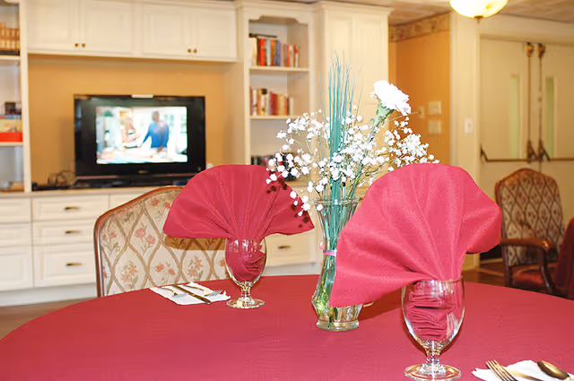 Round table set with a red tablecloth, folded red napkins in glasses and a floral centerpiece, with built-in cabinets and a TV in the background.