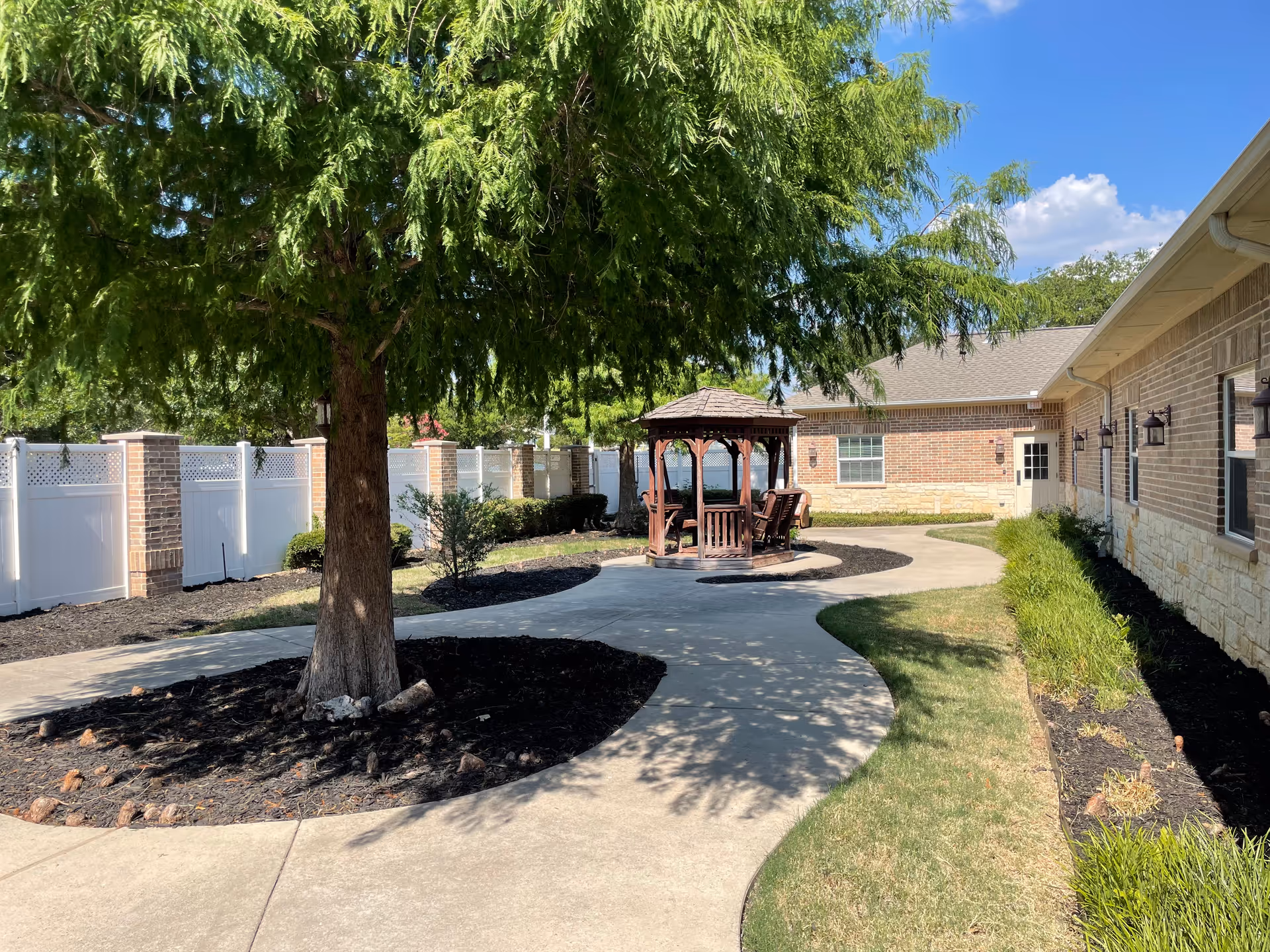 Outdoor garden area at Magnolia Assisted Living and Memory Care featuring a large tree with lush green foliage, a curved concrete pathway, a wooden gazebo with chairs underneath, and a brick building with windows and outdoor lights. The area is surrounded by a white fence and landscaped with grass and plants.