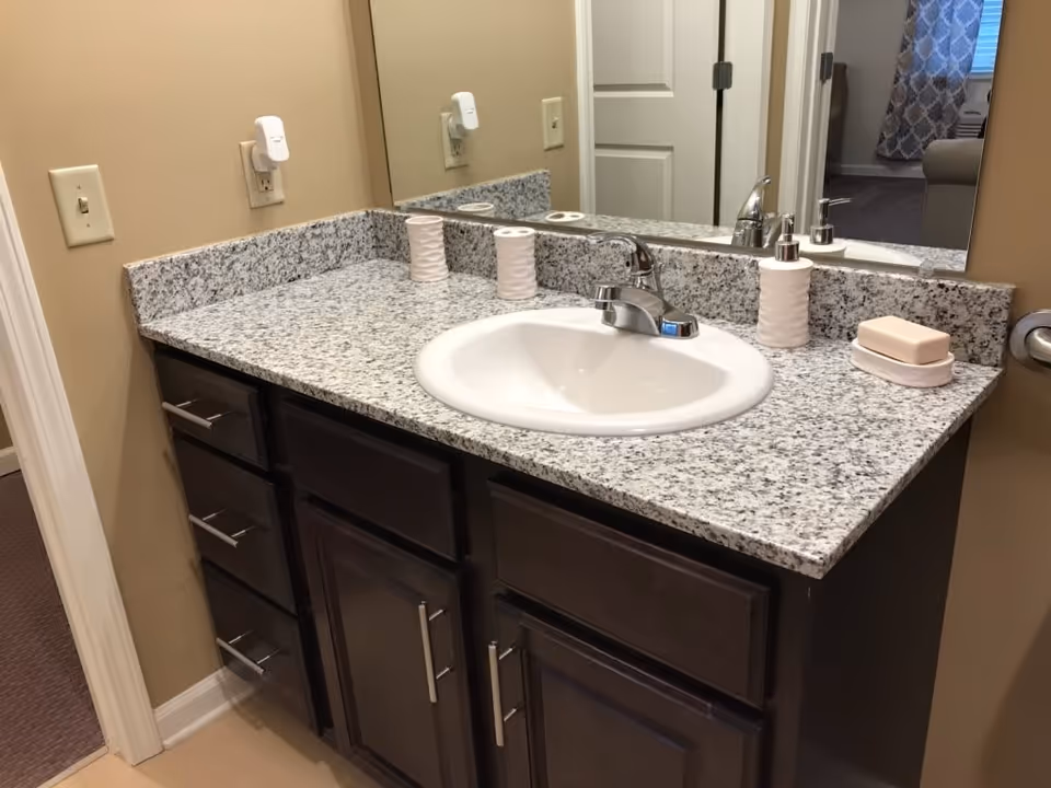 Bathroom vanity with a granite countertop, white sink, silver faucet, soap dispenser, soap dish with bar soap, and two white textured cups. There is a large mirror behind the sink reflecting part of the room, beige walls, and dark wood cabinets below the countertop.