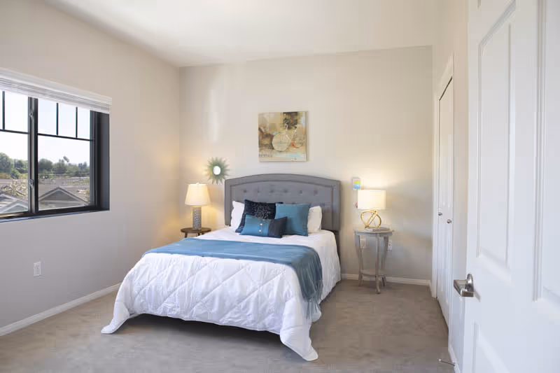 Bright, minimally decorated bedroom with a bed, upholstered headboard, two bedside tables with lamps, wall art, a window, and a closed door.