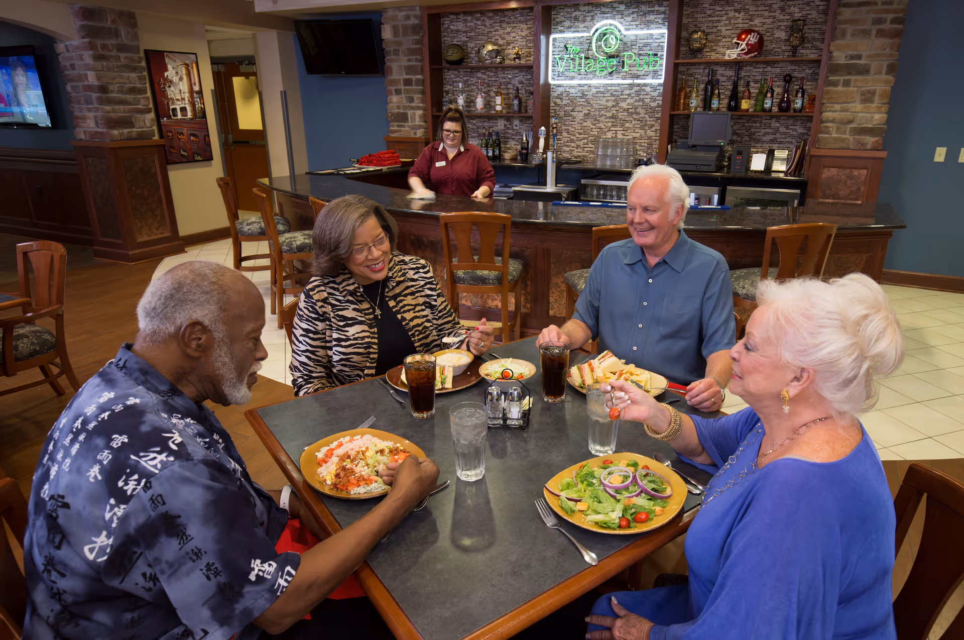 Four seniors eat and chat at a table in a pub-style dining area with a bartender and a neon 'Village Pub' sign behind the bar.