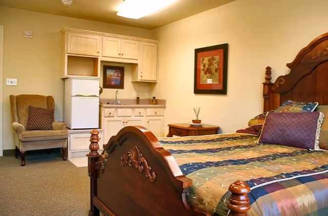 Furnished bedroom with an ornate wooden bed, colorful bedding, an armchair, and a small kitchenette with cabinets and a mini-fridge.