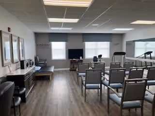Interior view of a senior living facility common area with rows of chairs facing a television and exercise equipment near large windows. The room has wood flooring, a bench along one wall, and framed pictures on the opposite wall.