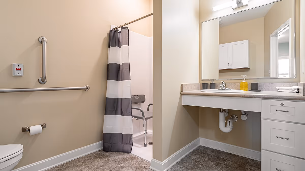 Accessible bathroom with beige walls, grab bars, a shower chair and striped curtain, and a sink with a large mirror.