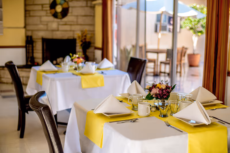 A dining area with tables covered in white tablecloths and yellow runners, each set with folded white napkins, plates, cups, glasses, and silverware. There are dark brown chairs around the tables, a stone fireplace in the background, and a glass door leading to an outdoor patio with tables and umbrellas.