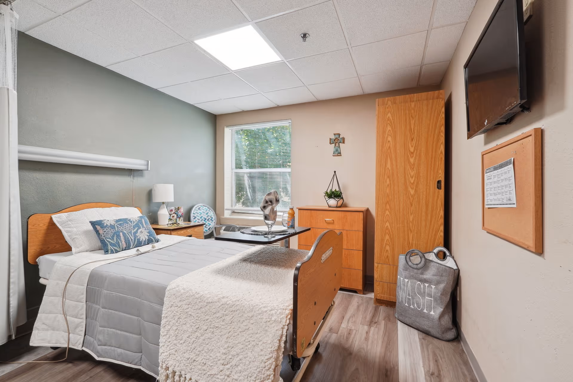 A neatly arranged nursing home bedroom with a single bed featuring a gray quilt and white blanket. There is a wooden headboard, a bedside table with a lamp and decorative items, a window with blinds, a wooden dresser with a small plant and cross on the wall above it, a mounted TV, a corkboard with a schedule, and a gray laundry bag labeled 'WASH' on the floor.